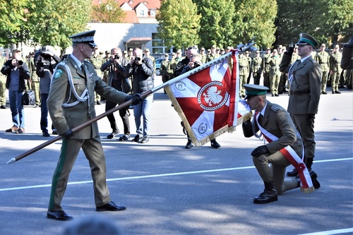 Rektor-Komendant pochyla sztandar przed sztandarowym, który klęka na prawe kolano, unosi płat do ust i całuje.